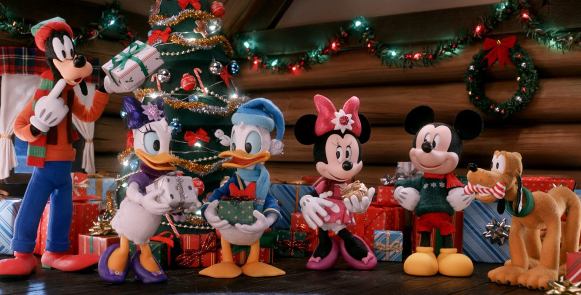 Standing in a log cabin are five stop-motion Disney characters, all dressed in Christmas attire and, except for Mickey Mouse, holding a wrapped Christmas present. Pictured from left to right are Goofy, Daisy Duck, Donald Duck, Minnie Mouse, Mickey Mouse, and Pluto. Behind them, more wrapped gifts are sitting on the floor beneath a decorated Christmas tree and a log wall decorated with a lighted garland and a lighted wreath.