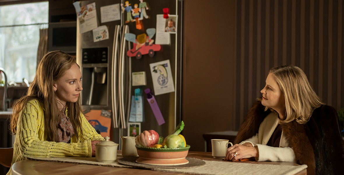 In an image from FX’s Fargo: Year 5, Dorothy “Dot” Lyon (Juno Temple) is sitting at a kitchen table talking with another woman (Jennifer Jason Leigh). Dot is wearing a yellow sweater over a blouse; the other woman is wearing a brown fur coat and a beige suit jacket. They each have a coffee cup in front of them, and there is a refrigerator behind them, covered in magnets and children’s art.