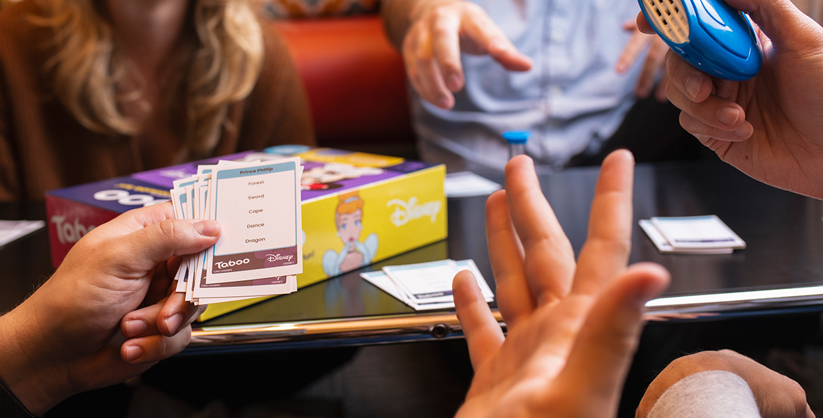 Four people sit around a black table, playing DL2_0577.jpg. In the foreground of the image, the hands of a player are extended, holding some of the game’s cards. Beside this person is another hand, holding the game’s blue buzzer. In the background, two more players smile and point at the players in the foreground.