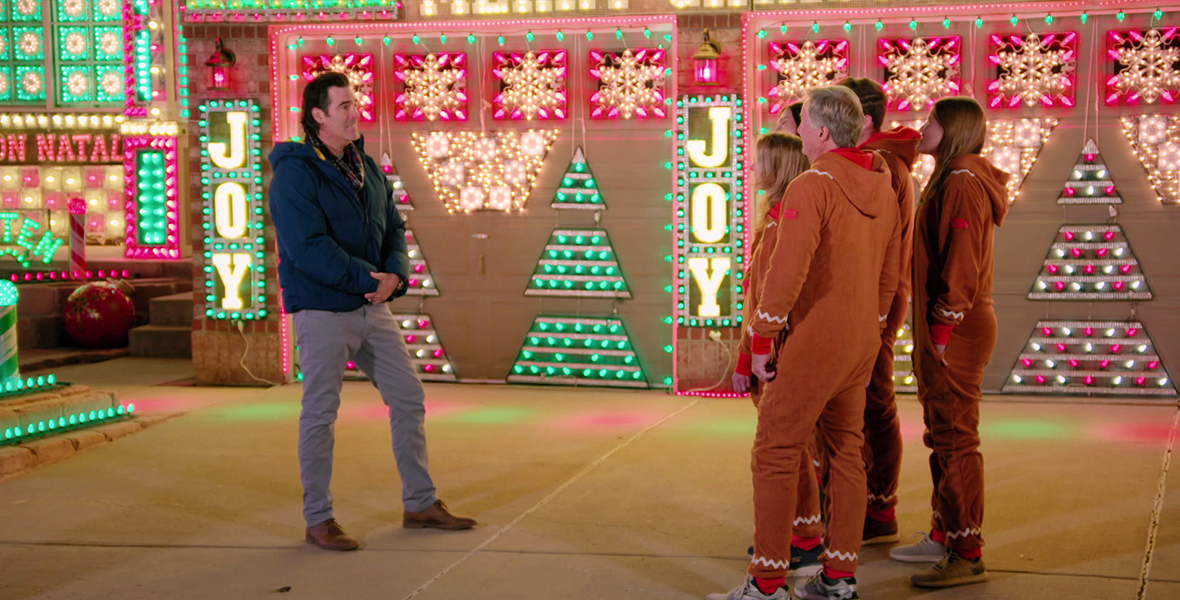 In an image from ABC’s The Great Christmas Light Fight, host/judge Carter Oosterhouse stands outside one of the houses being judged on Sunday’s episode. He stands to the left of the family, who are all wearing reindeer onesies; Oosterhouse is wearing a blue jacket and gray pants. Plentiful sparking Christmas lights are attached to the house and the garage doors seen behind them.